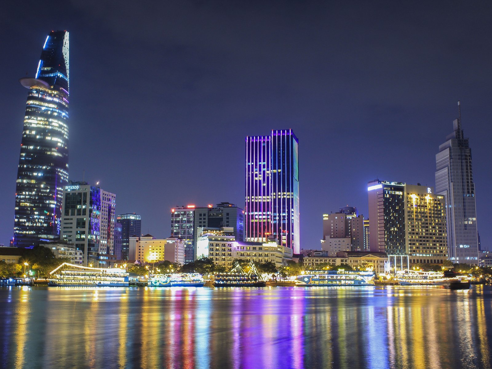 Saigon riverfront promenade at dusk, with a tree-lined avenue and city towers along the water
